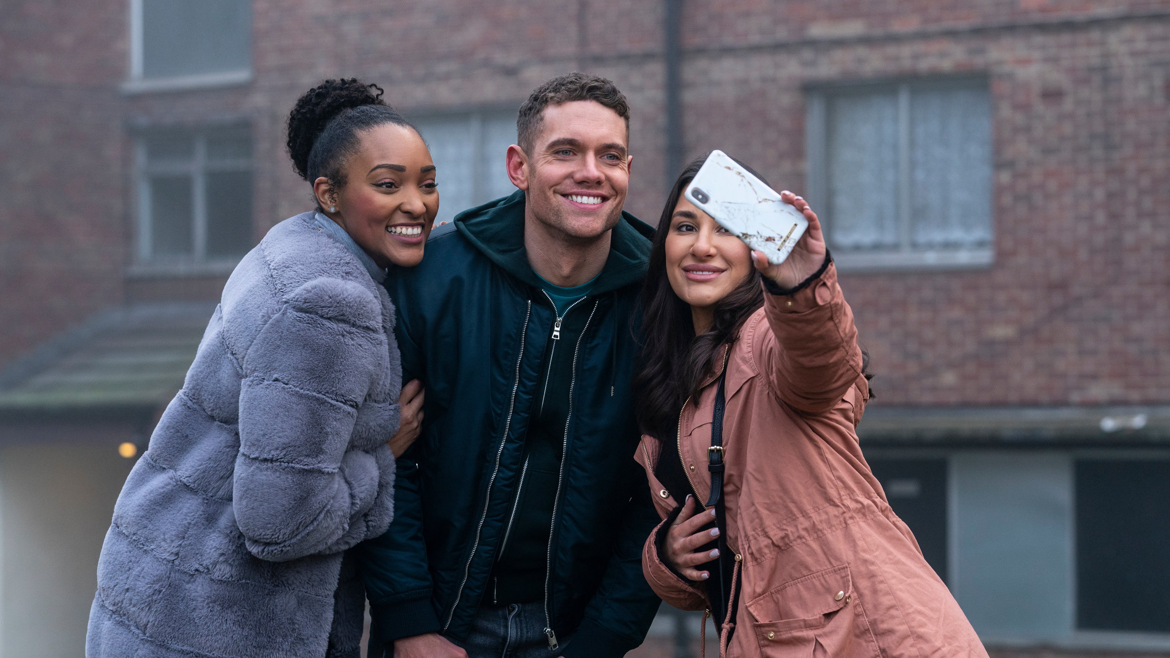 "Make Me Famous": Billy (Tom Brittney) steht zwischen zwei Frauen, von denen eine ein Handy hochhält, um ein Selfie zu machen. Alle lächeln in die Kamera.