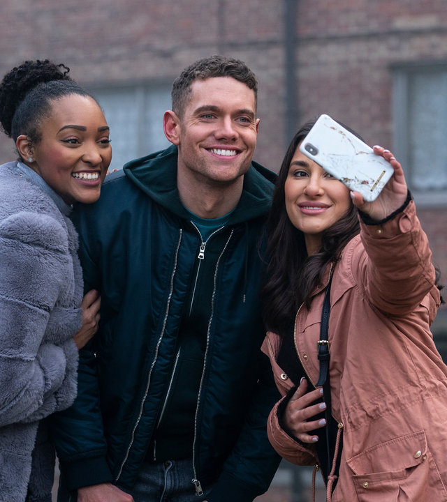 "Make Me Famous": Billy (Tom Brittney) steht zwischen zwei Frauen, von denen eine ein Handy hochhält, um ein Selfie zu machen. Alle lächeln in die Kamera.
