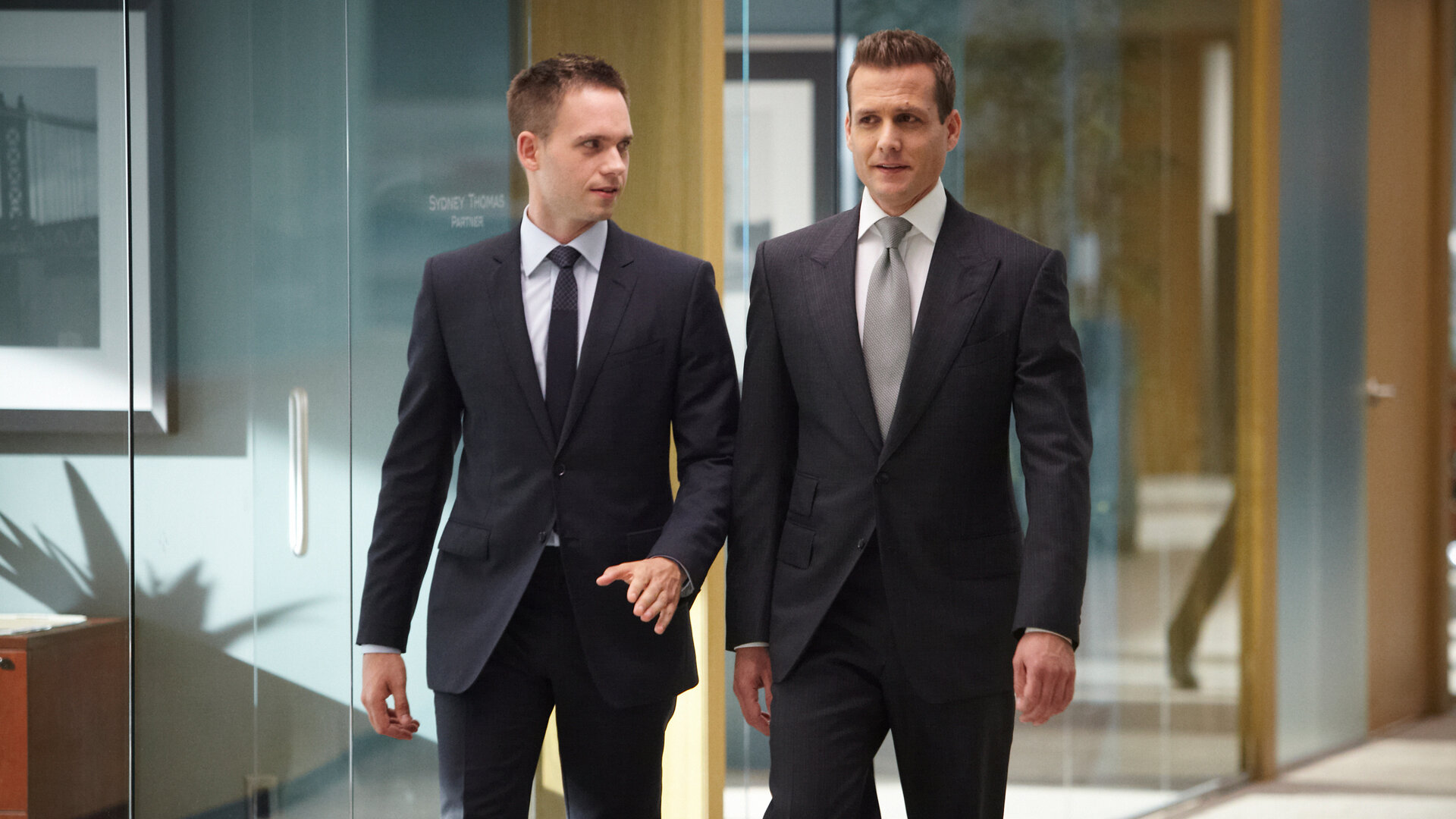 "Suits - Verlieren, um zu gewinnen": Mike (Patrick J. Adams) und Harvey (Gabriel Macht) gehen nebeneinander durch einen gläsernen Büroflur. Beide tragen dunkle Anzüge; Mike schaut zu Harvey, während dieser geradeaus blickt.