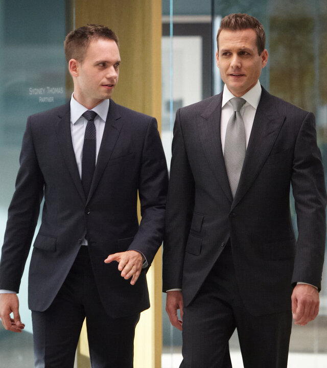 "Suits - Verlieren, um zu gewinnen": Mike (Patrick J. Adams) und Harvey (Gabriel Macht) gehen nebeneinander durch einen gläsernen Büroflur. Beide tragen dunkle Anzüge; Mike schaut zu Harvey, während dieser geradeaus blickt.