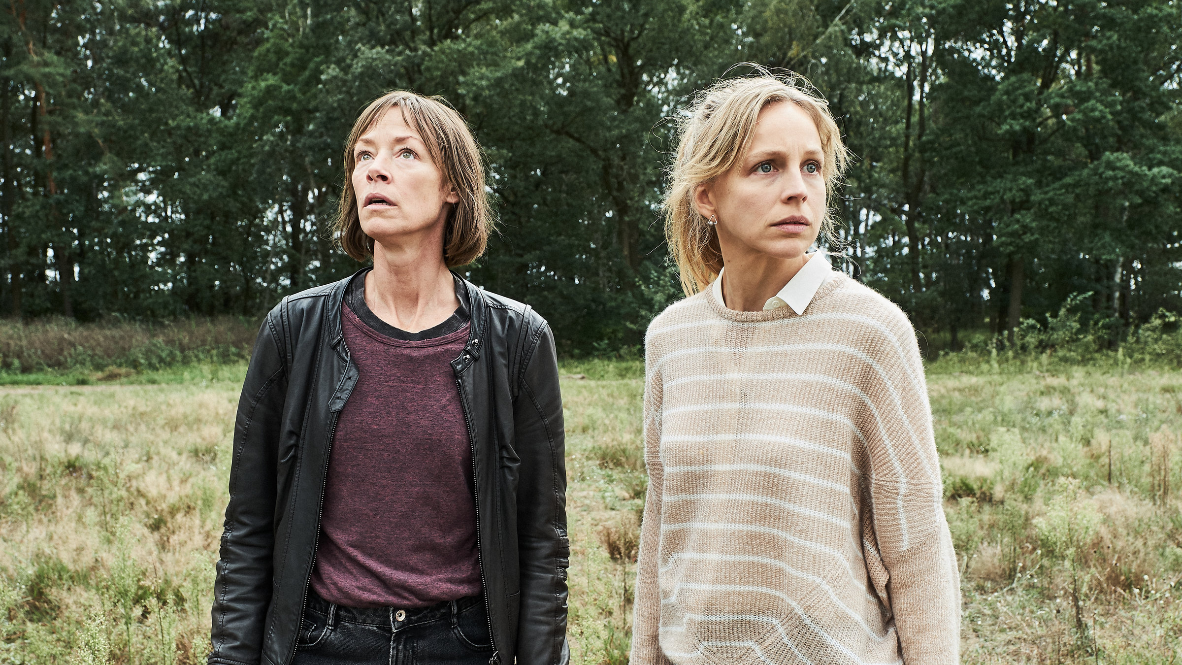 "Der Mordanschlag (2)": Bettina Polheim (Jenny Schily) und Sandra Wellmann (Petra Schmidt-Schaller) stehen auf einer Wiese. Im Hintergrund ist ein kleines Wäldchen zu sehen. Bettina sieht sehr aufmerksam nach oben, Sandra schaut ängstlich um sich.
