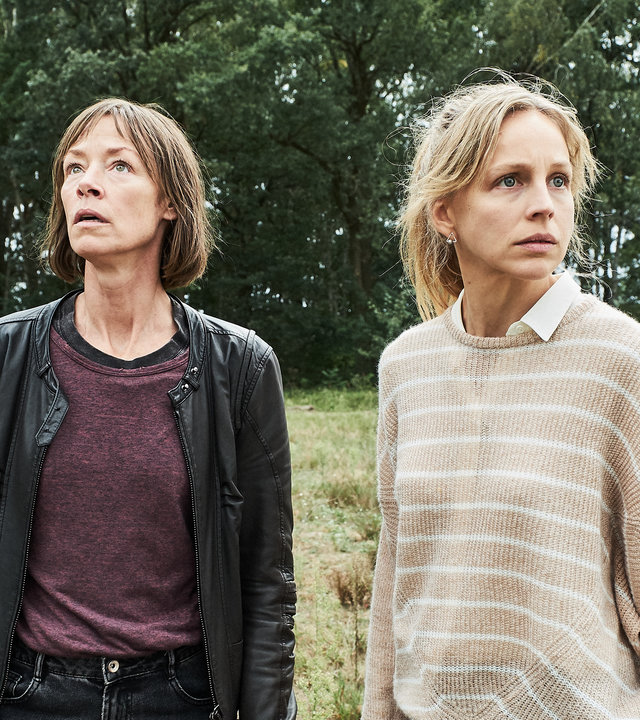 "Der Mordanschlag (2)": Bettina Polheim (Jenny Schily) und Sandra Wellmann (Petra Schmidt-Schaller) stehen auf einer Wiese. Im Hintergrund ist ein kleines Wäldchen zu sehen. Bettina sieht sehr aufmerksam nach oben, Sandra schaut ängstlich um sich.