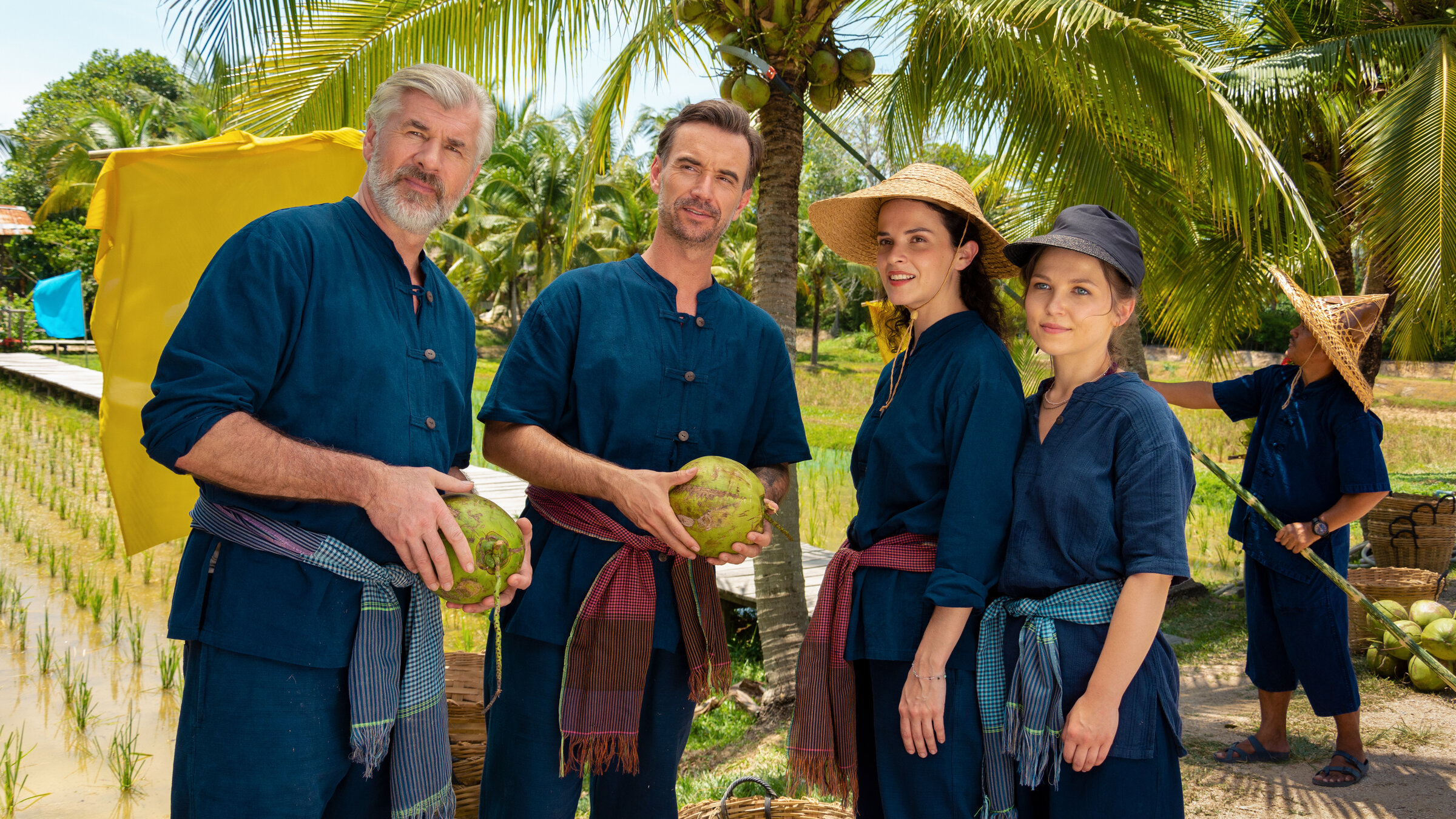 "Das Traumschiff - Phuket": Auf einer Kokosnussplantage: Max Parger (Florian Silbereisen), Martin Grimm (Daniel Morgenroth), Annika Ehler (Maxine Kazis) und Melly Sturm (Marija Mauer) stehen zusammen und blicken in die Ferne. Sie tragen traditionelle Bauernkleidung und die Frauen Hüte. Die Männer halten Kokosnüsse in den Händen.