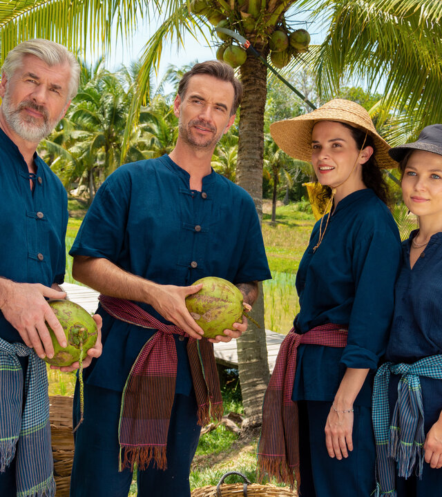 "Das Traumschiff - Phuket": Auf einer Kokosnussplantage: Max Parger (Florian Silbereisen), Martin Grimm (Daniel Morgenroth), Annika Ehler (Maxine Kazis) und Melly Sturm (Marija Mauer) stehen zusammen und blicken in die Ferne. Sie tragen traditionelle Bauernkleidung und die Frauen Hüte. Die Männer halten Kokosnüsse in den Händen.