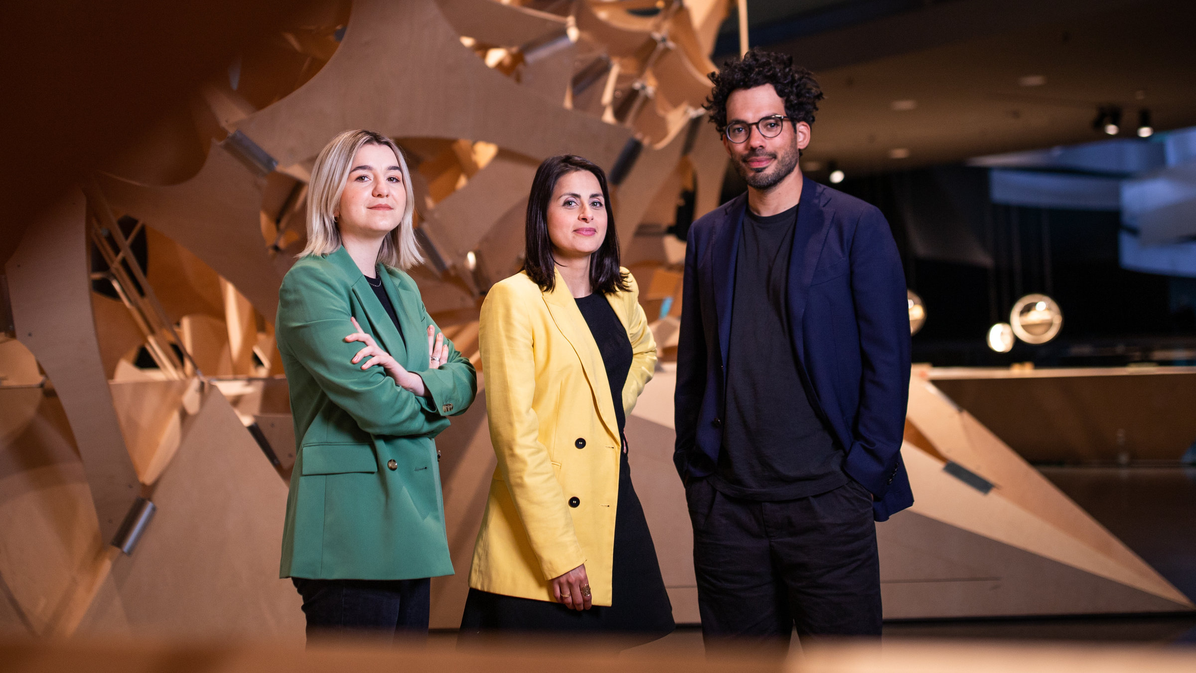 "Futur Wir – Das neue deutsche Selbstverständnis (2/3) - Wie wir lieben wollen": Die drei Moderatoren stehen im Berliner Futurium und blicken in die Kamera. Eingerahmt sind sie von einer großen Holzskulptur.
