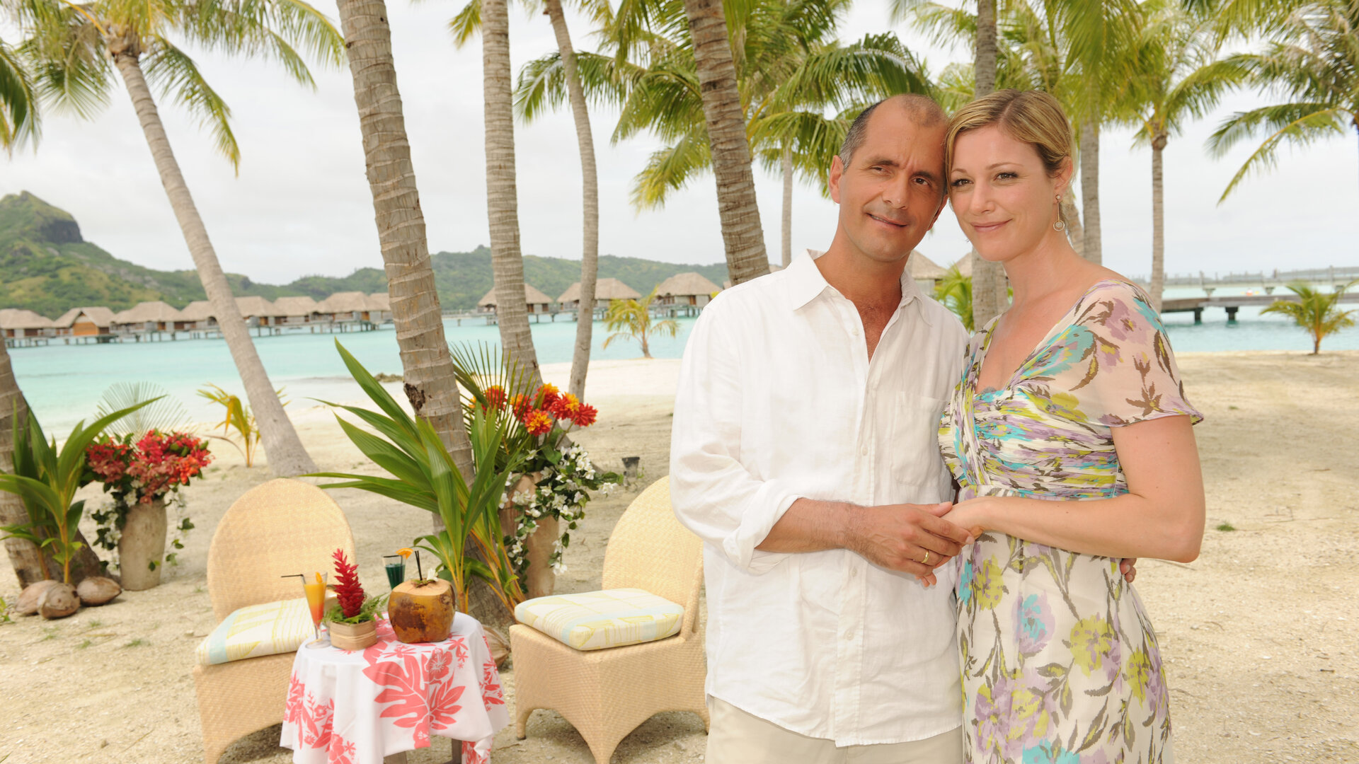 "Das Traumschiff: Bora Bora": Sabine (Julia Stinshoff) und ihr Mann Marek (Christoph Maria Herbst) stehen lächelnd Arm in Arm am Strand. Im Hintergrund ein mit Cocktails gedeckter Tisch.