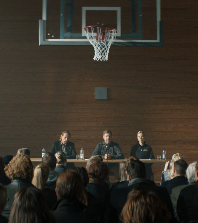 "Cryptid - Die falsche Todesursache": Der Schulleiter Axel (Johan Hedenberg) und die Polizistin Petra (Angelina Håkansson) sitzen in der Schulturnhalle einer Menge Menschen gegenüber.