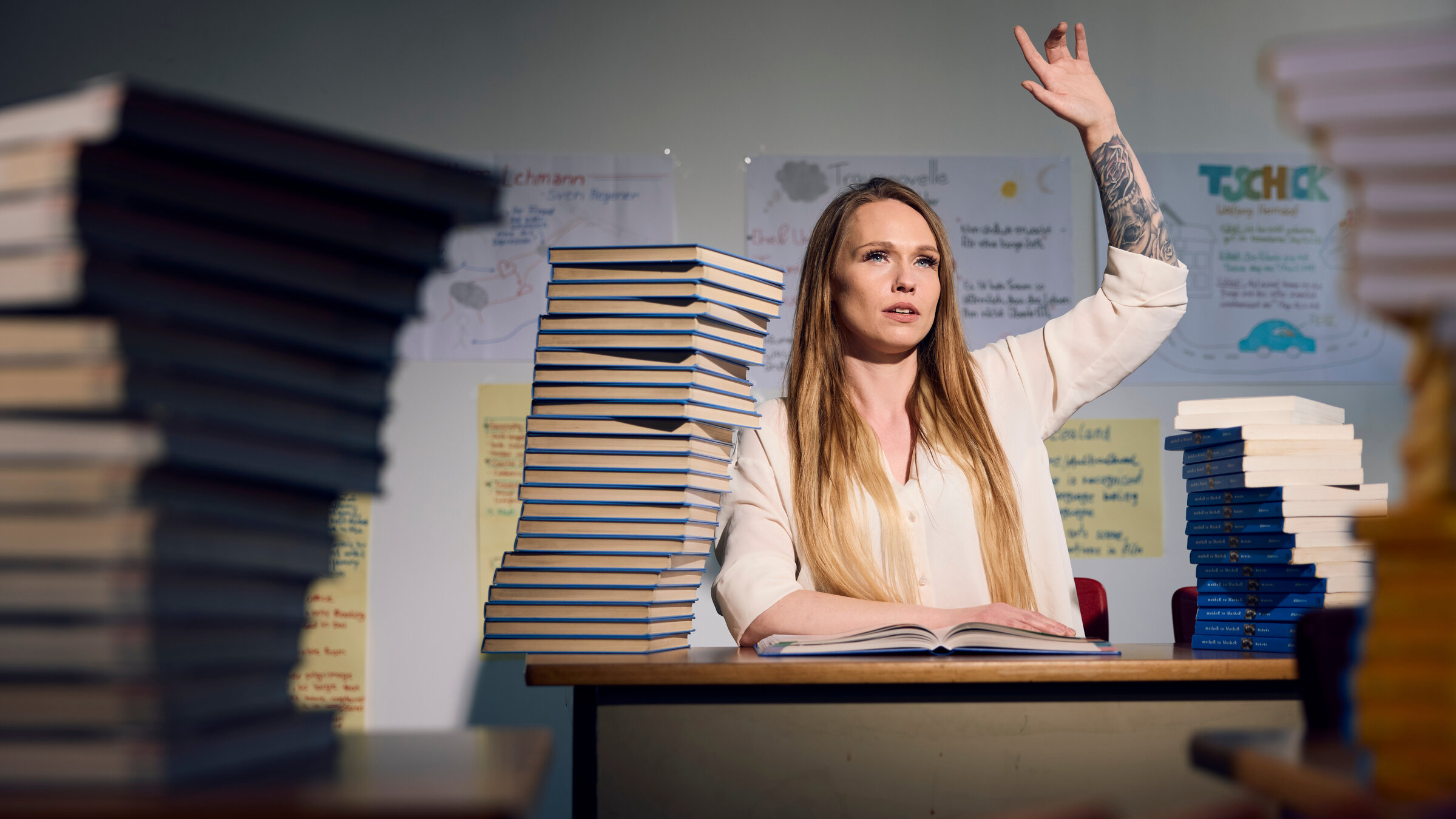 "37°Leben - Lebensretter Schule: Jeder Tag ein Kampf": Sarah sitzt auf einer typischen Schulbank. Vor ihr liegt ein Berg an Schulbüchern. Sie meldet sich, augenscheinlich um eine Antwort zu geben. Im Hintergrund sind Plakate mit Lerninhalten zu erkennen.
