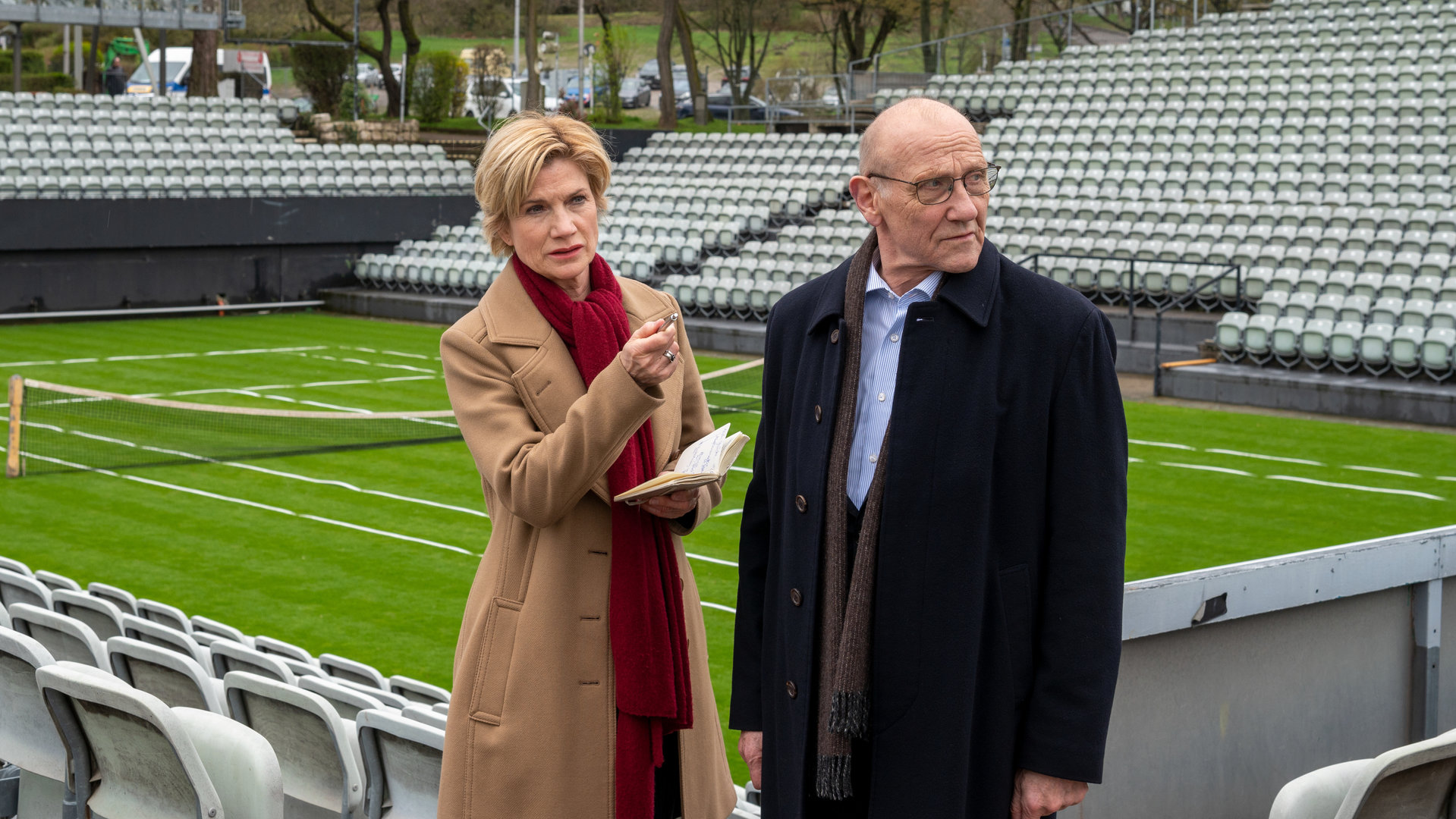 "SOKO Stuttgart - K.o. im zweiten Satz": Kriminalhauptkommissarin Martina Seiffert (Astrid M. Fünderich) steht neben Kriminaldirektor Michael Kaiser (Karl Kranzkowski) auf der Zuschauertribüne eines Tennisplatzes. Sie hält ein Notizbuch in der Hand, mit der anderen Hand zeigt sie in die Ferne. Martina trägt einen braunen Mantel und einen roten Schal. Der Kriminaldirektor, ebenfalls mit Schal und dunkler Jacke bekleidet, schaut sich um.