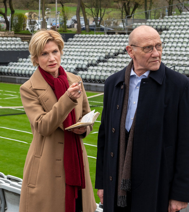 "SOKO Stuttgart - K.o. im zweiten Satz": Kriminalhauptkommissarin Martina Seiffert (Astrid M. Fünderich) steht neben Kriminaldirektor Michael Kaiser (Karl Kranzkowski) auf der Zuschauertribüne eines Tennisplatzes. Sie hält ein Notizbuch in der Hand, mit der anderen Hand zeigt sie in die Ferne. Martina trägt einen braunen Mantel und einen roten Schal. Der Kriminaldirektor, ebenfalls mit Schal und dunkler Jacke bekleidet, schaut sich um.