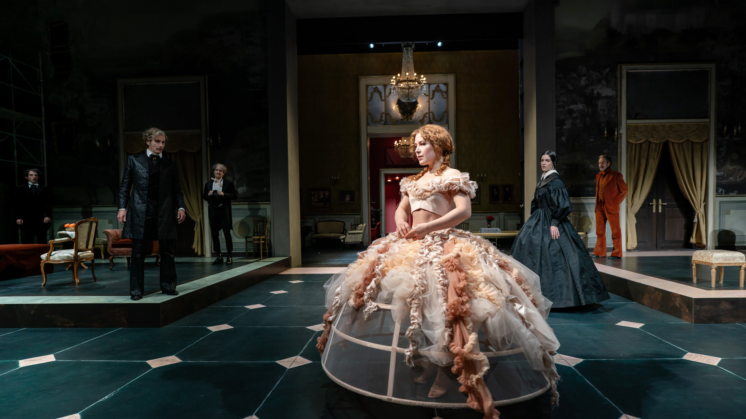 "Il Gattopardo": Große Bühnenaufstellung mit einer Frau in einem rosafarbenen, weit ausgestellten Reifrockkleid und roten, gelockten Haaren im Vordergrund; weitere Personen stehen verteilt im Raum eines klassizistischen Salons.