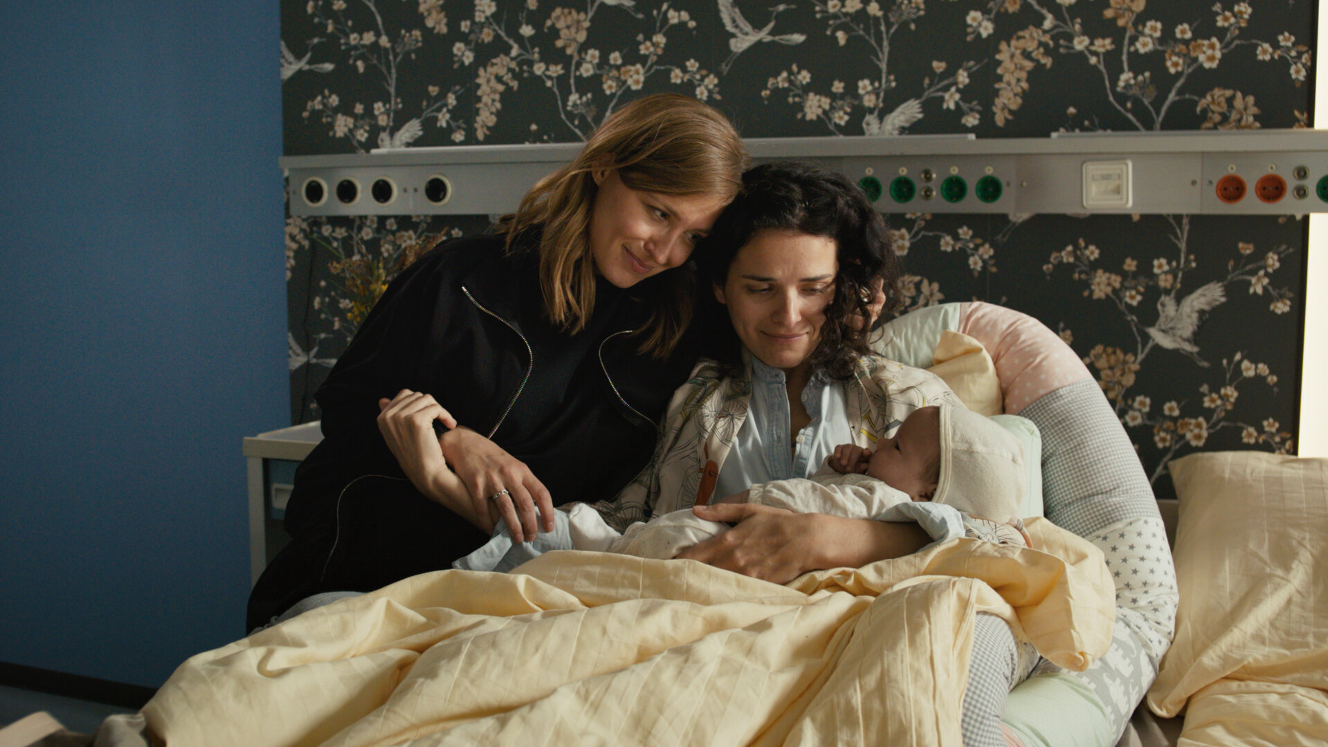 "Lena Lorenz - Kreuz und queer": In einem Krankenhausbett sitzt Franzi Winkler-Ertl (Maxine Kazis) und hält lächelnd ein Baby im Arm. Alex Winkler-Ertl (Pina Kühr) sitzt neben ihr, schmiegt sich an sie und betrachtet lächelnd das Baby.