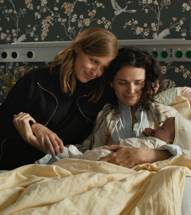 "Lena Lorenz - Kreuz und queer": In einem Krankenhausbett sitzt Franzi Winkler-Ertl (Maxine Kazis) und hält lächelnd ein Baby im Arm. Alex Winkler-Ertl (Pina Kühr) sitzt neben ihr, schmiegt sich an sie und betrachtet lächelnd das Baby.
