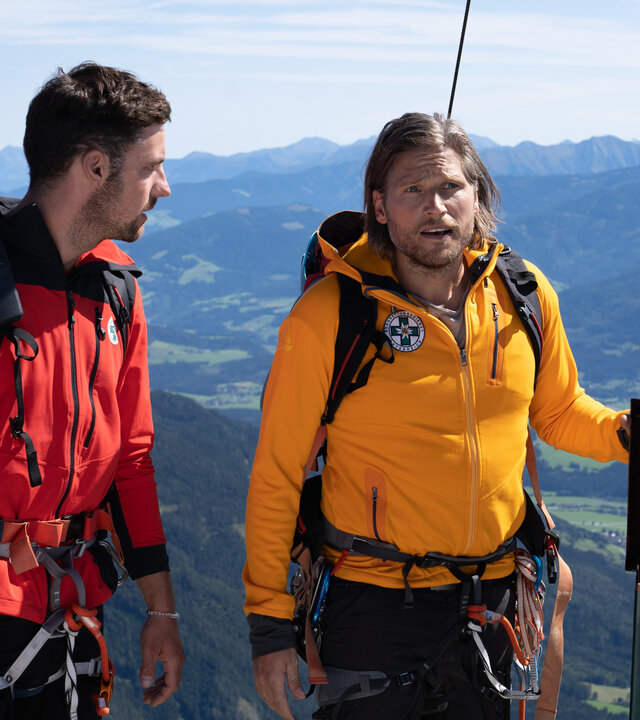"Die Bergretter: Im Gipfelbuch (2)": Die Bergretter Markus (Sebastian Ströbel) und Simon (Ferdinand Seebacher) stehen am Gipfel eines Berges und wirken überrascht. Markus hält mit einer Hand eine Klappe fest, die normalerweise das Gipfelbuch sichert.