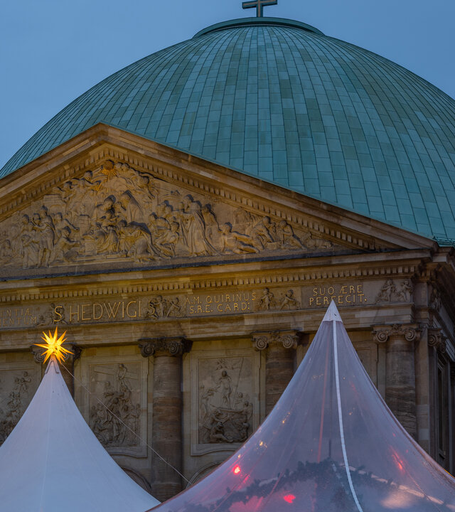 "Katholischer Gottesdienst - Freude im Herzen, Hoffnung im Blick": Außenansicht der St. Hedwigs-Kathedrale in Berlin mit Weihnachtsständen im Vordergrund.