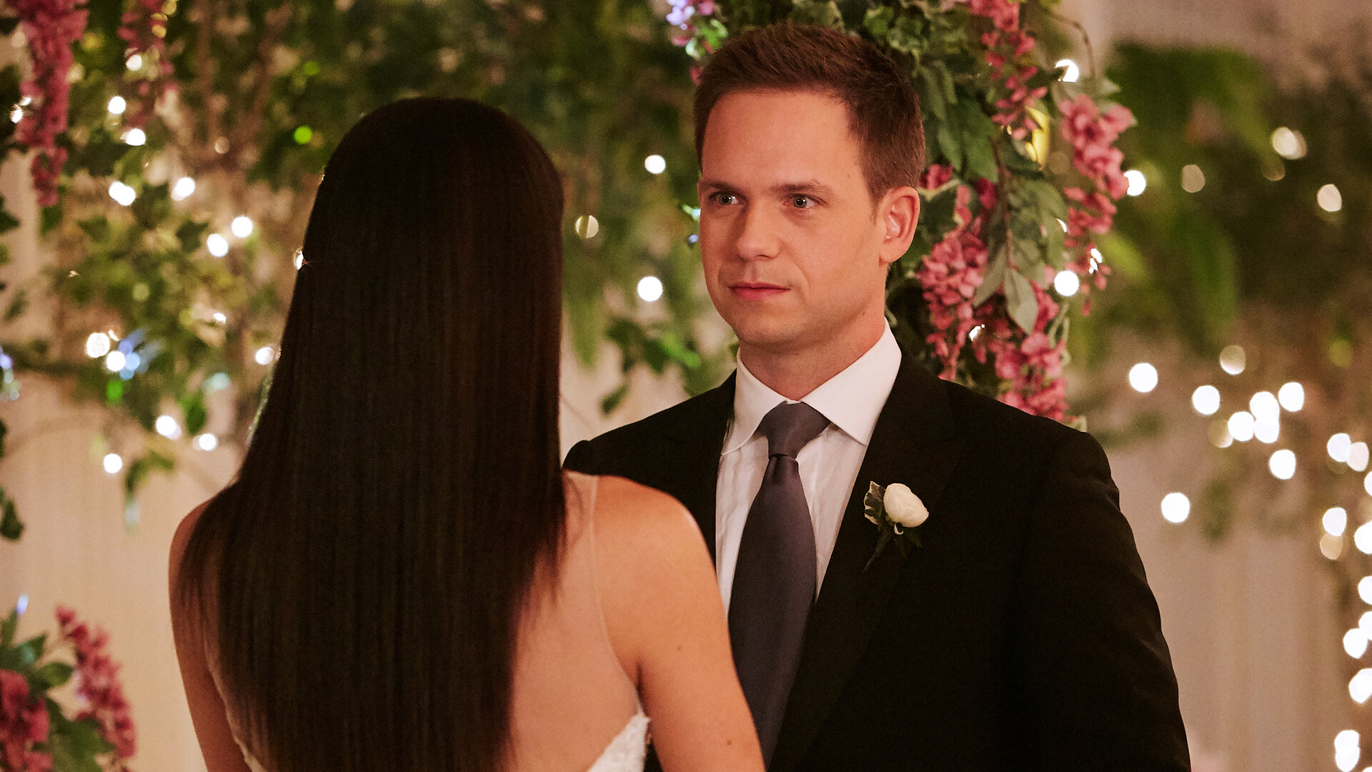 "Suits - Abschied": Mike (Patrick J. Adams) steht im Hochzeitsanzug unter einem kunstvollen Blumenarrangement, das mit Lichterketten versehen eine feierliche Atmosphäre verbreitet, Rachel (Meghan Markle) gegenüber, deren Rücken der Kamera zugewandt ist.