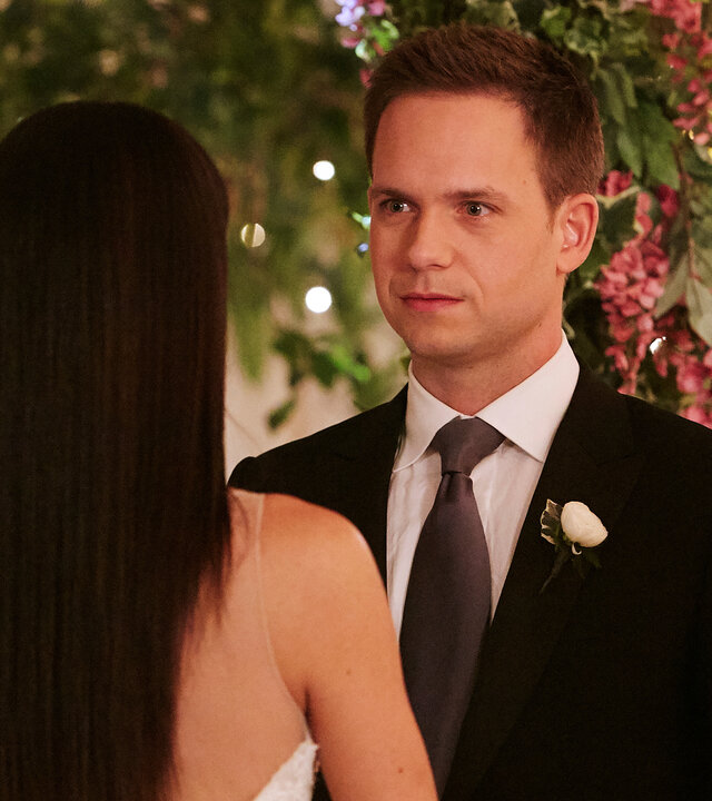 "Suits - Abschied": Mike (Patrick J. Adams) steht im Hochzeitsanzug unter einem kunstvollen Blumenarrangement, das mit Lichterketten versehen eine feierliche Atmosphäre verbreitet, Rachel (Meghan Markle) gegenüber, deren Rücken der Kamera zugewandt ist.