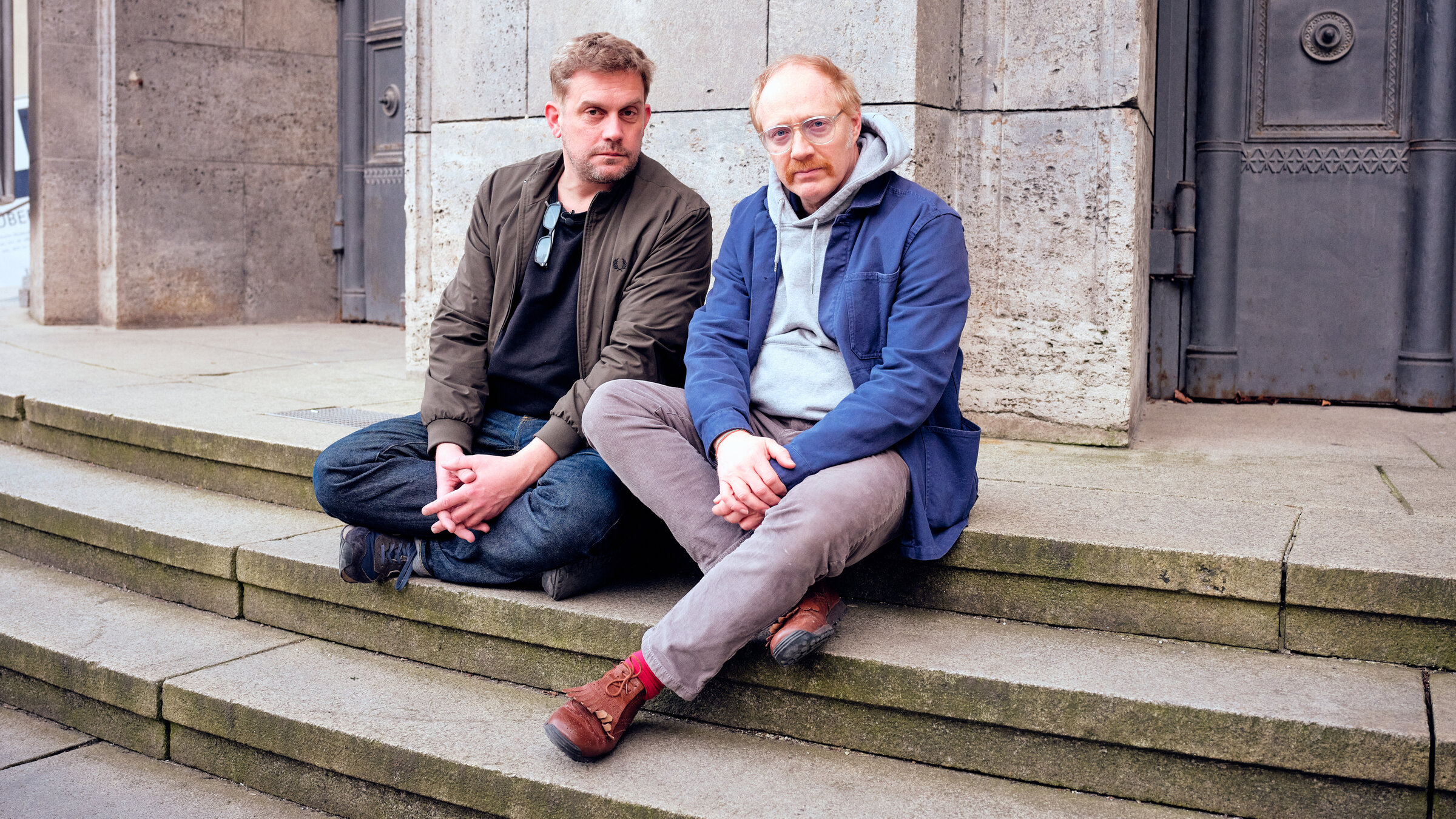 "Bezzel & Schwarz - Die Grenzgänger - Im Museum": Die "Grenzgänger" Sebastian Bezzel (links) und Simon Schwarz sitzen auf den Stufen vor dem Eingang des Deutschen Museums.