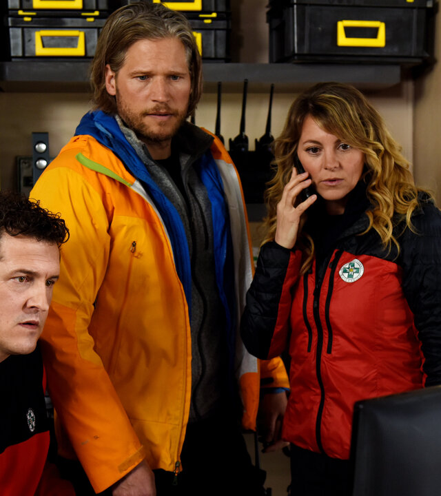 "Die Bergretter - Dünnes Eis (1)": In der Bergretter-Zentrale: Bergretter Rudi (Michael Pascher) sitzt vor einem Monitor. Sein Kollege Markus (Sebastian Ströbel) steht, wie auch Bergretterin Katharina (Luise Bähr) neben ihm. Sie telefoniert mit dem Handy. Mit entsetzten Blicken starren sie auf den Bildschirm.