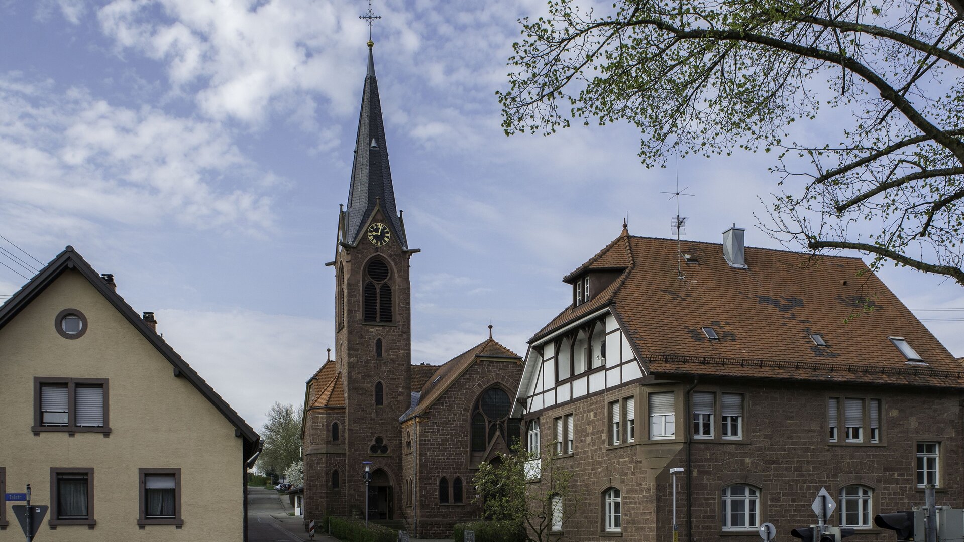 "Evangelischer Gottesdienst - Aufstehen": Außenansicht der Waldenserkirche in Palmbach, daneben stehen zwei Häuser der Altstadt und an der Seite befinden sich die Äste eines Baumes. Der Kirchturm ragt in den leicht bewölkten Himmel.