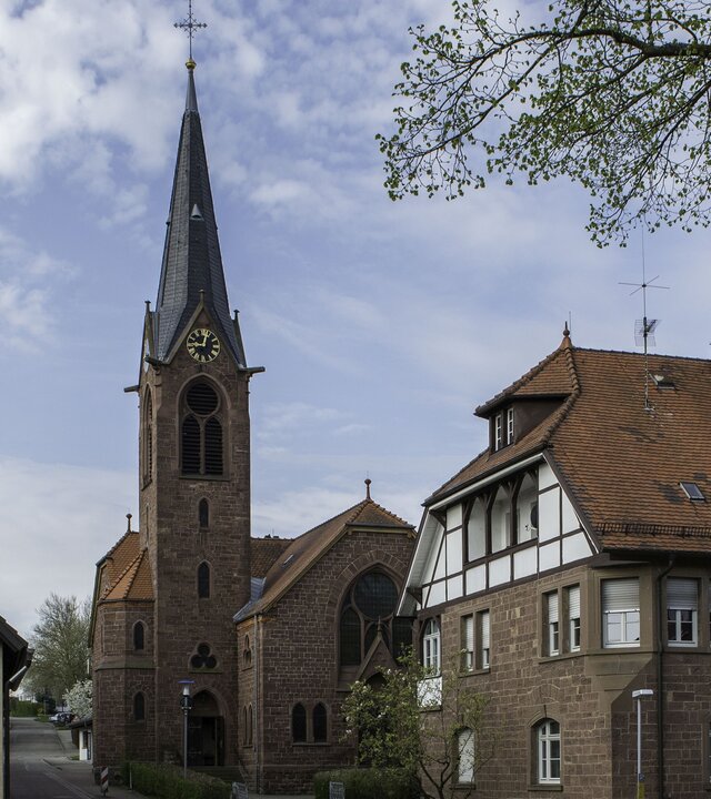 "Evangelischer Gottesdienst - Aufstehen": Außenansicht der Waldenserkirche in Palmbach, daneben stehen zwei Häuser der Altstadt und an der Seite befinden sich die Äste eines Baumes. Der Kirchturm ragt in den leicht bewölkten Himmel.