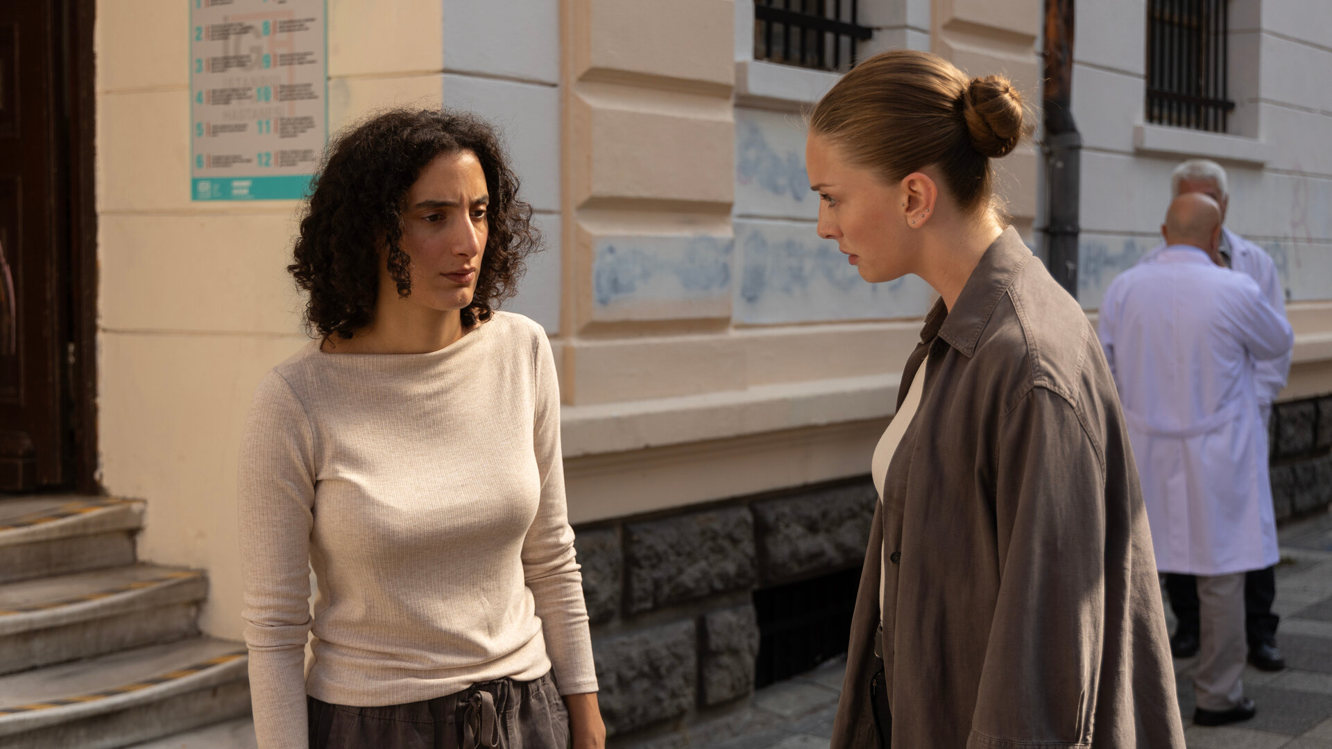 "Inspector Ikmen - Tod in Istanbul: Die Stadt und der Müll (2)": Tagsüber im Sonnenschein auf einer Straße: Leah (Mouna Soualem) und Ayse (Yasemin Kay Allen) stehen sich vor einem opulenten Gebäude mit vergitterten Fenstern gegenüber und blicken sich ernst an.