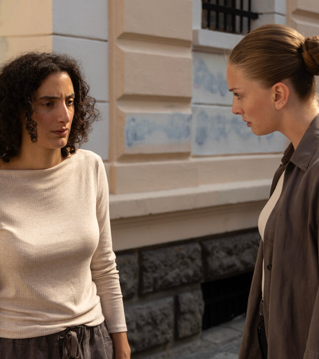 "Inspector Ikmen - Tod in Istanbul: Die Stadt und der Müll (2)": Tagsüber im Sonnenschein auf einer Straße: Leah (Mouna Soualem) und Ayse (Yasemin Kay Allen) stehen sich vor einem opulenten Gebäude mit vergitterten Fenstern gegenüber und blicken sich ernst an.