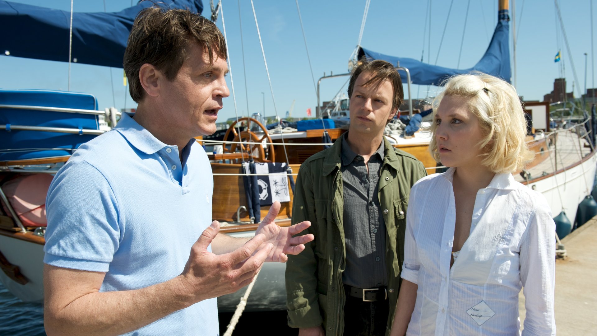 "SOKO Wismar - Zerbrochenes Glas": Kommissar Herzog (Michael Härle) steht mit Magnus (Tobias Hoesl) und Annika Norlin (Nikola Kastner) auf einem Bootsanleger im Jachthafen. Hinter ihnen ist ein Segelboot mit blauen Segeln zu erkennen.