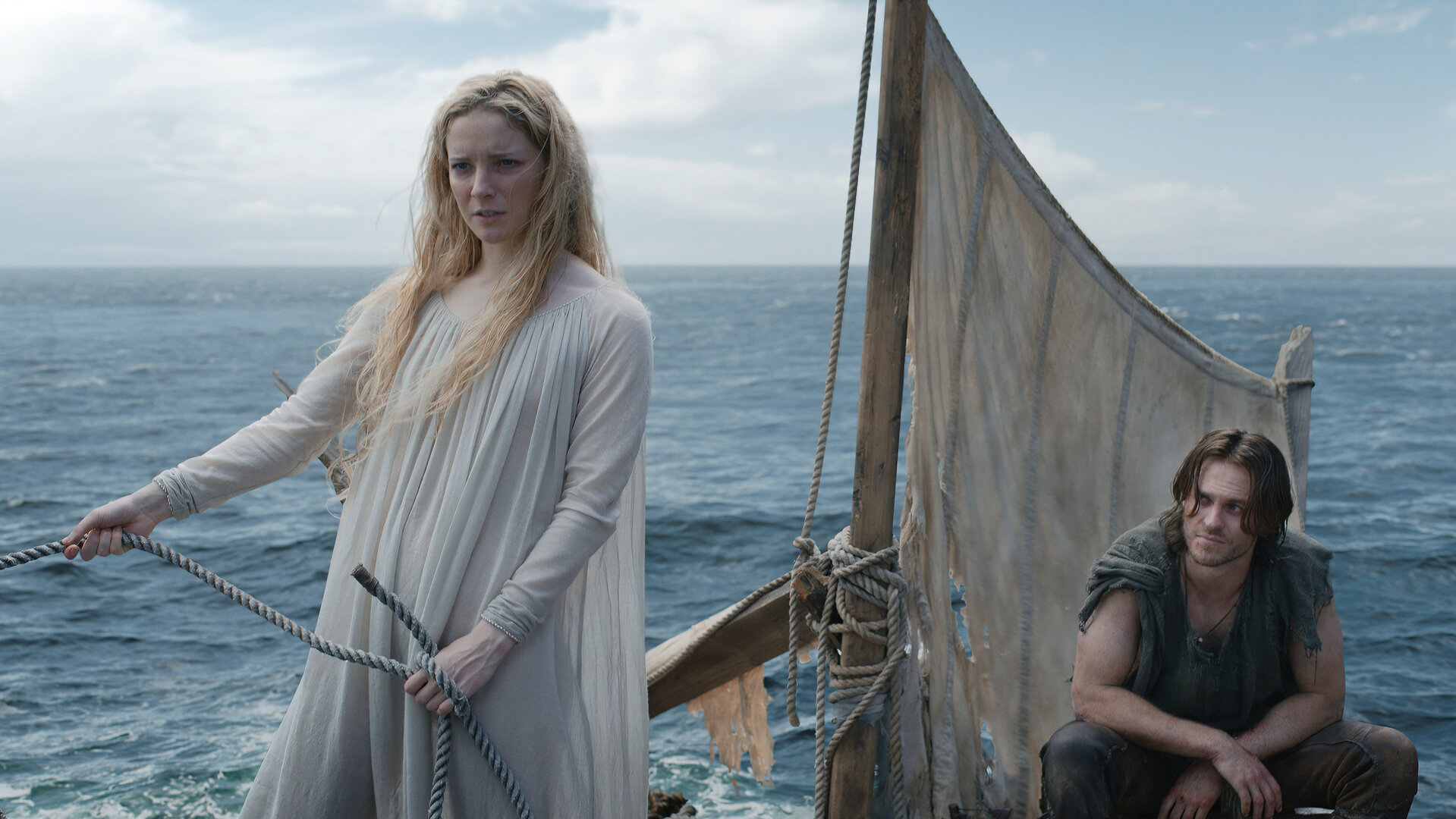 "Der Herr der Ringe: Die Ringe der Macht - Treibgut": Galadriel (Morfydd Clark) steht auf einem Floß mit einem kleinen Segel. Hinter ihr hockt Halbrand (Charlie Vickers). Im Hintergrund ist das Meer zu sehen.