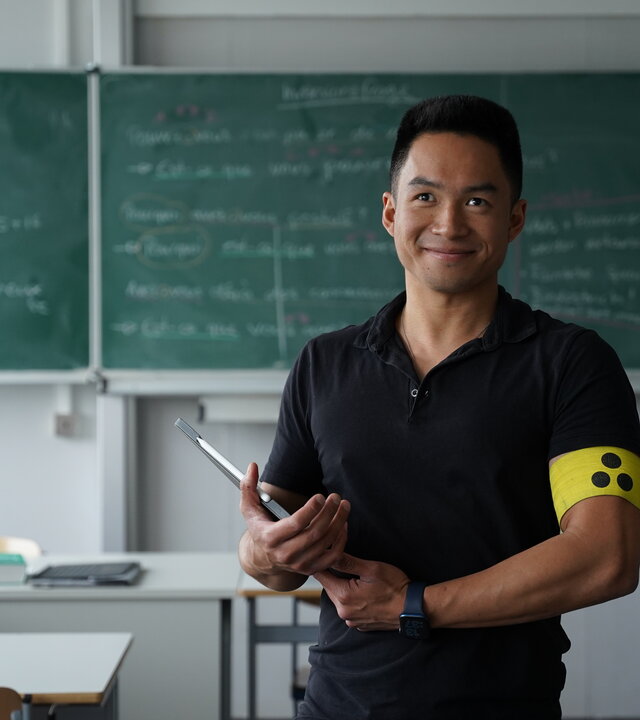 "einfach Mensch: Wai Long Van - Von Vielen unterschätzt (1)": Wai Long Van steht mit einer gelben Armbinde mit drei schwarzen Punkten vor einer Schultafel. Er blickt lächelnd nach vorne.