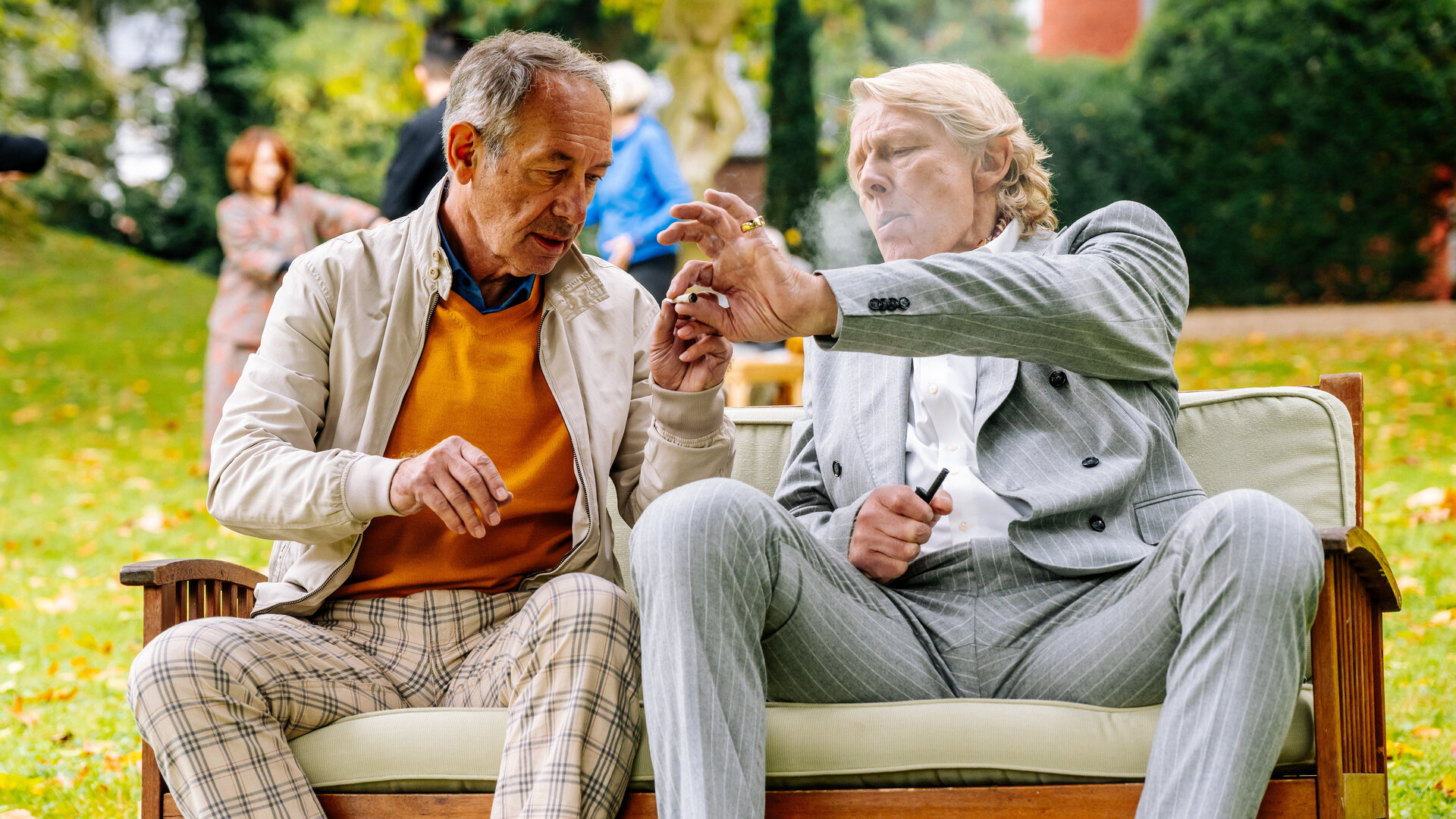 "SOKO Köln - Forever Young": Carl Henke (Alexander von der Groeben) und Richard Bartz (Robert Hunger-Bühler) sitzen nebeneinander auf einer Holzbank auf einer Wiese. Carl Henke pustet Rauch aus und reicht Richard Bartz eine angefangene Zigarette. Im Hintergrund sind verschwommen drei Personen (Komparsen) auf der Wiese zu erkennen.