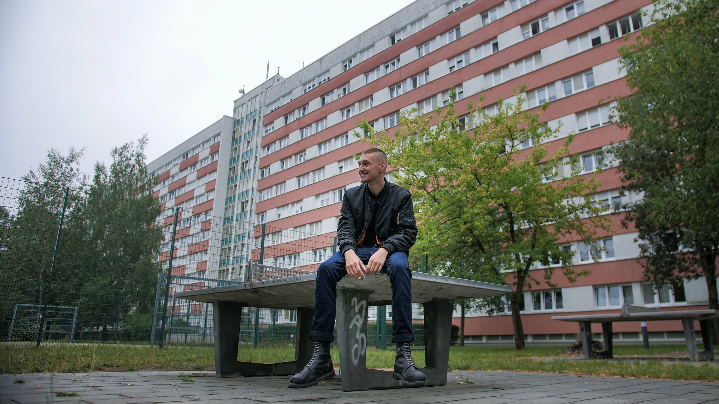 "37°: 333 Meter Hausflur – Leben in der "Langen Lene"": Paul Abend sitzt auf einer Tischtennisplatte und schaut lächelnd links an der Kamera vorbei. Im Hintergrund sind Bäume und ein rot-weißer Plattenbau zu sehen.