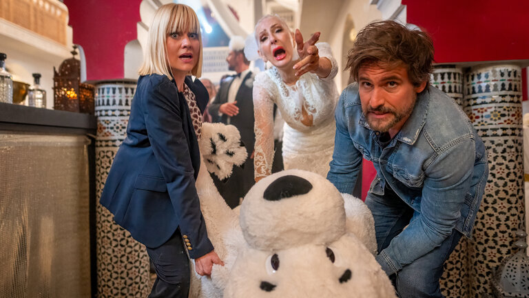 "Keine Scheidung ohne Leiche": Nina (Henrike Fehrs) und Tom (Tom Beck) ziehen mit beiden Händen ein Eisbärkostüm über den Boden. Sie befinden sich in einer Bar im marokkanischen Stil, auf der Seite stehen Getränkeflaschen auf einem Tresen. Hinter den beiden steht Juliane (Desirée Nick) in ihrem Hochzeitskleid. Sie beugt sich nach vorne und streckt ihren linken Arm aus und ruft entsetzt. Im Hintergrund sieht man noch Gäste (Komparsen) der Hochzeitsfeier.