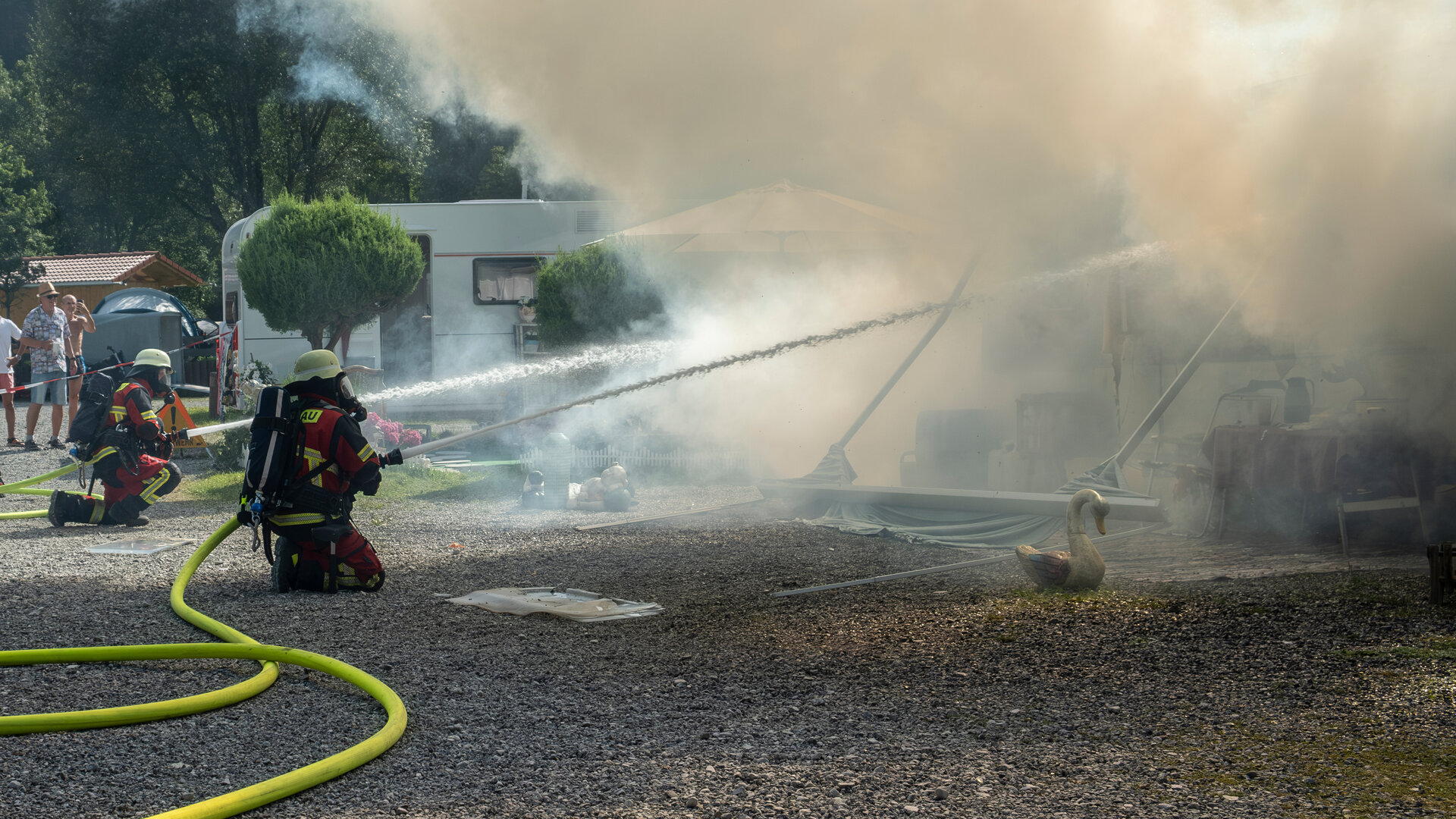 "Marie fängt Feuer - Vergeben und Vergessen": Marie (Christine Eixenberger) kniet zusammen mit einem Kollegen der Feuerwehr auf dem Boden des Campingplatzes Beide zielen mit Feuerwehrschläuchen auf brennende Wohnwagen, die sich hinter einer Rauchwolke befinden.