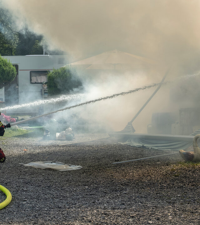 "Marie fängt Feuer - Vergeben und Vergessen": Marie (Christine Eixenberger) kniet zusammen mit einem Kollegen der Feuerwehr auf dem Boden des Campingplatzes Beide zielen mit Feuerwehrschläuchen auf brennende Wohnwagen, die sich hinter einer Rauchwolke befinden.
