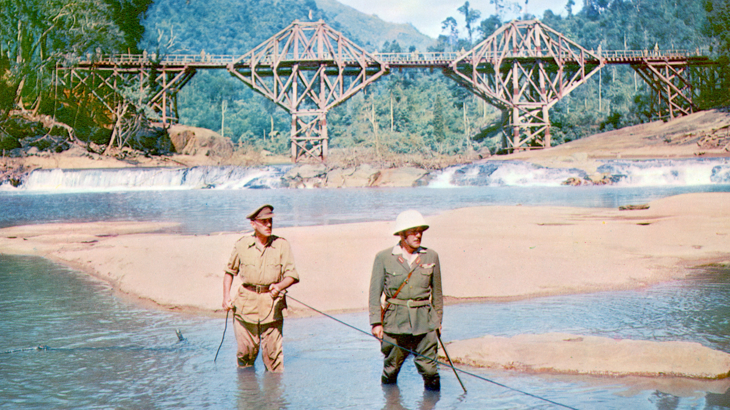 "Die Brücke am Kwai": Im Vordergrund stehen zwei uniformierte Männer knietief im Wasser eines tropischen Flusses. Colonel Nicholson (Alec Guiness, links) hält ein Kabel in der Hand, das rechts aus dem Bild herausführt. Im Hintergrund ist vor der Kulisse bewaldeter Hügel die imposante Holzkonstruktion einer Eisenbahnbrücke zu sehen.