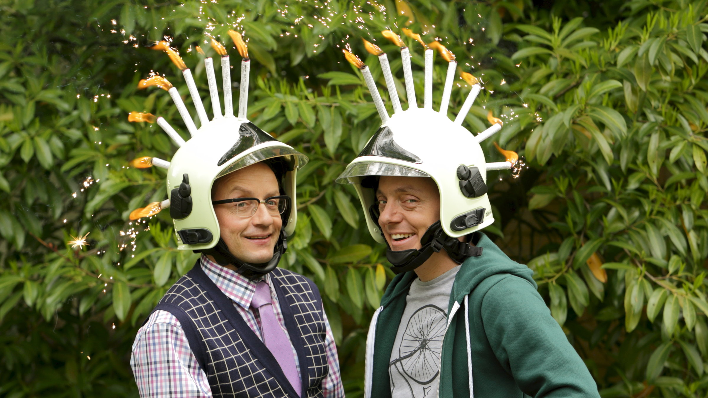 "Nicht nachmachen!":  Bernhard Hoëcker und Wigald Boning stehen vor einer Hecke. Beide tragen Helme, auf denen Feuerwerk brennt.