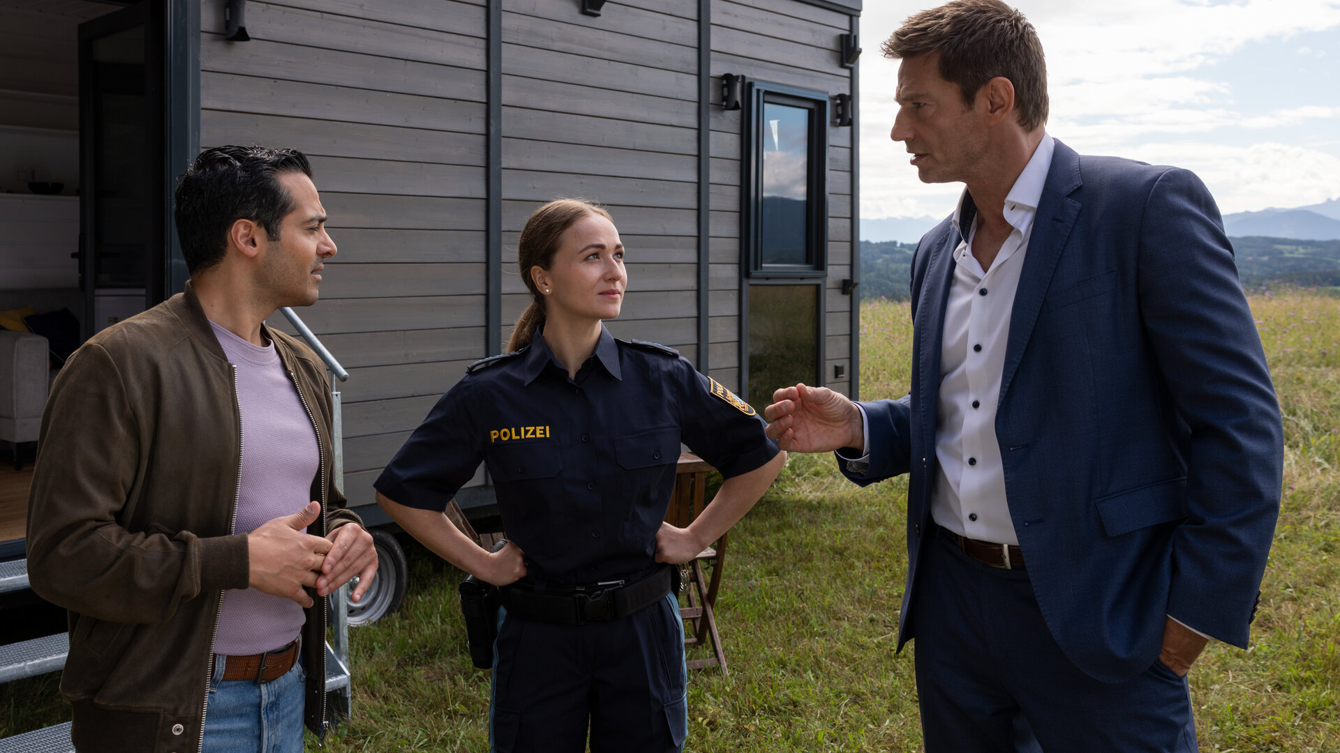"Die Rosenheim-Cops - Tod im Tiny House": Kommissar Kaya (Baran Hêvî), Polizistin Pia Arnbruck (Katharina Plank) und Hauptkommissar Hansen (Igor Jeftić) stehen auf einer Wiese vor einem dunkelbraunen Tiny House und sprechen miteinander.