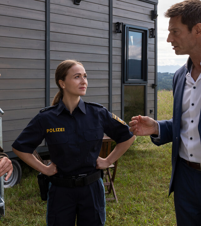 "Die Rosenheim-Cops - Tod im Tiny House": Kommissar Kaya (Baran Hêvî), Polizistin Pia Arnbruck (Katharina Plank) und Hauptkommissar Hansen (Igor Jeftić) stehen auf einer Wiese vor einem dunkelbraunen Tiny House und sprechen miteinander.