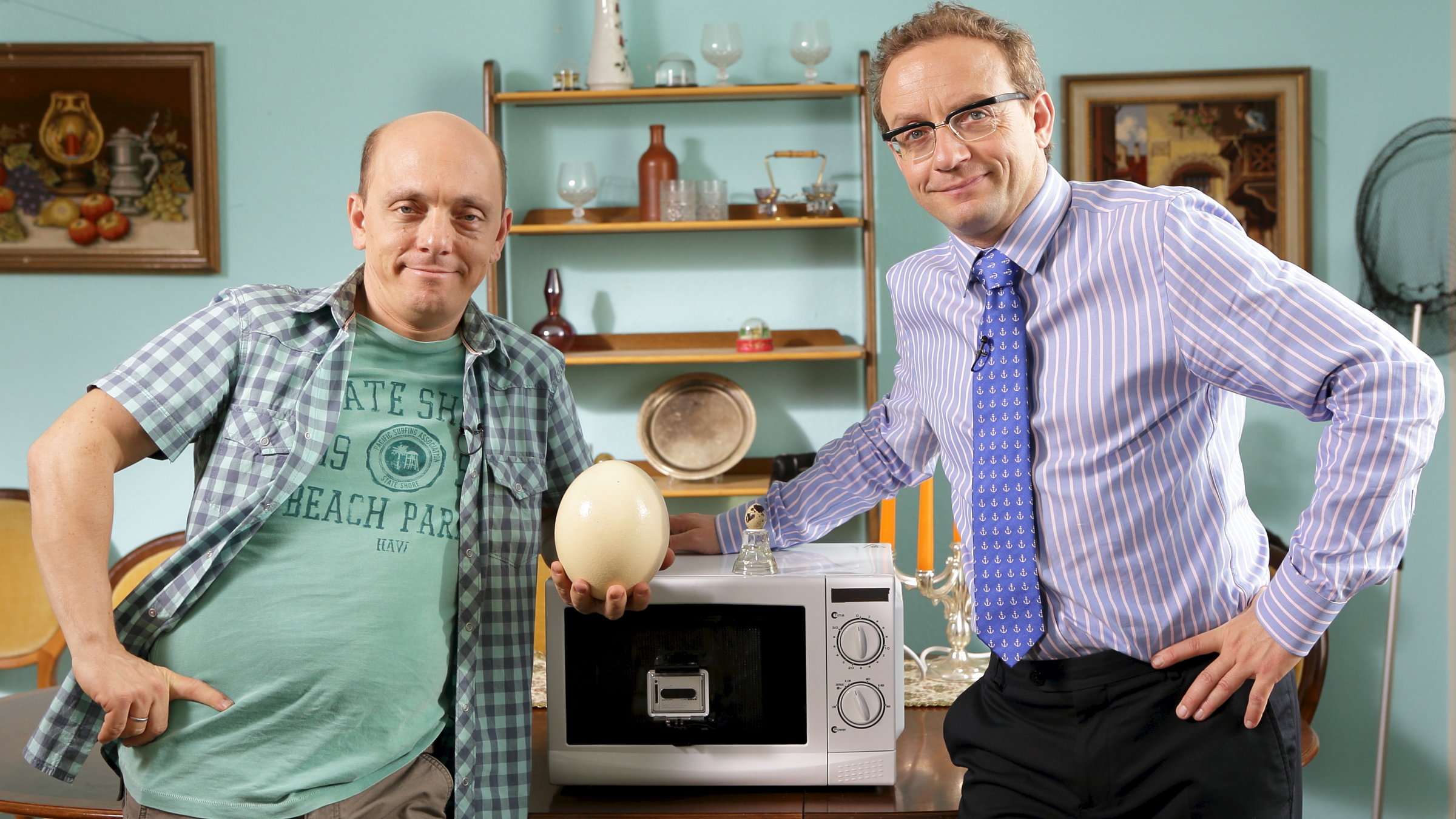 "Nicht Nachmachen": Bernhard Hoëcker und Wigald Boning stehen an einem Tisch, auf dem eine Mikrowelle steht. Beide sehen Richtung Kamera. Bernhard Hoëcker hält ein Straußenei in der Hand.