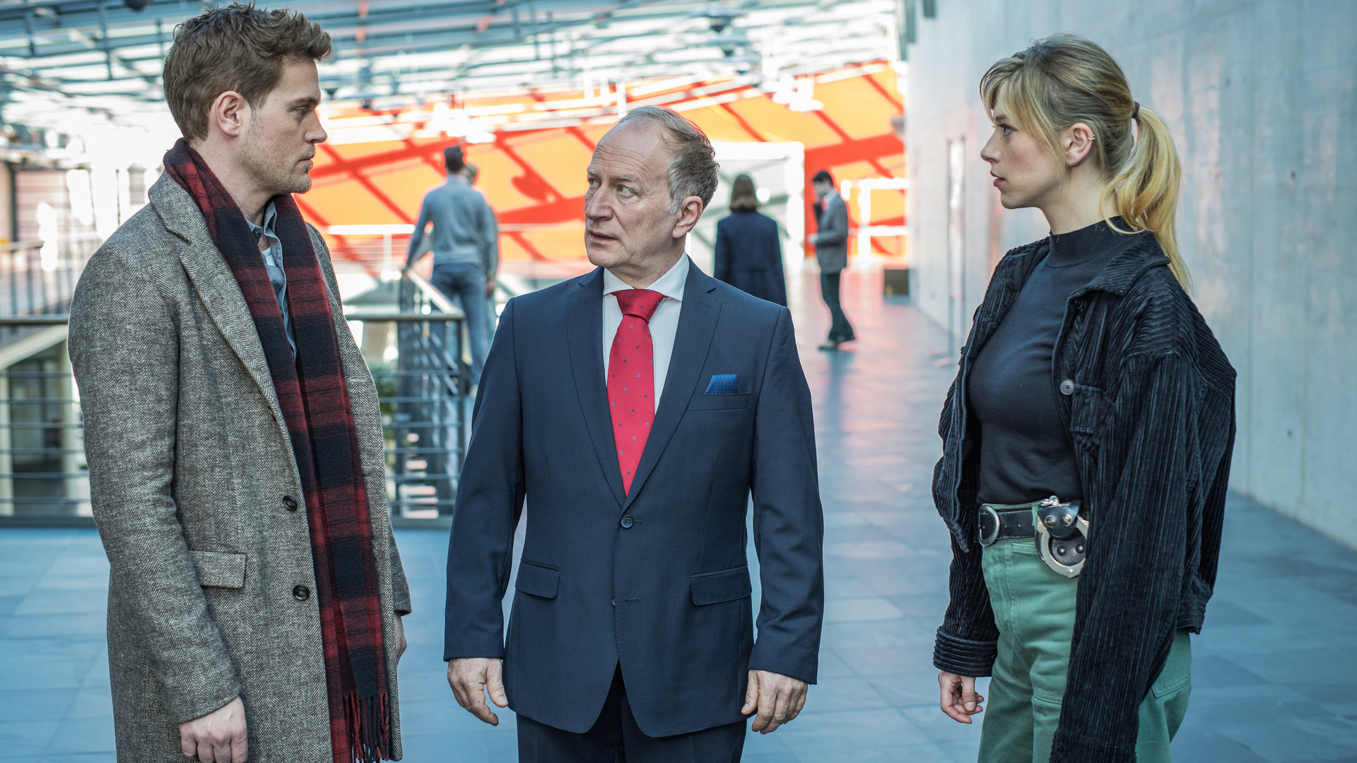 "SOKO München - Affenliebe": Die Kommissare Resi Schwaiger (Mersiha Husagic) und Dominik Morgenstern (Joscha Kiefer) stehen mit Gerhard Beck (Stefan Merki), dem Geschäftsführer des Institutes, an dem das Opfer studiert hat, im Flur. Beck trägt einen edlen Anzug und steht zwischen den beiden Kommissaren, während er Dominik Morgenstern entsetzt in die Augen schaut. Resi Schwaiger sieht währenddessen zu ihrem Kollegen.