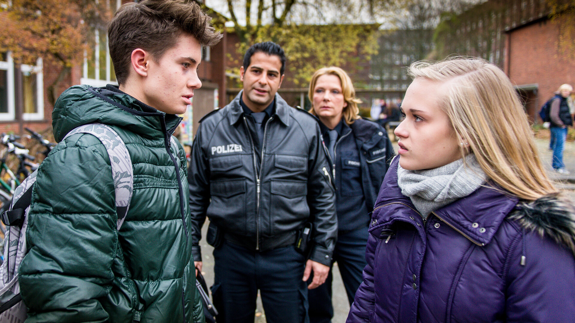 "Notruf Hafenkante - Verhinderte Liebe": Im Innenhof einer Schule stehen sich René Marwort (Marvin Jaacks) und Jette Lürsen (Nadine Kösters) gegenüber und schauen sich an. Im Hintergrund stehen die Polizisten Tarik Coban (Serhat Cokgezen) und Claudia Fischer (Janette Rauch), die die beiden Jugendlichen beobachten.