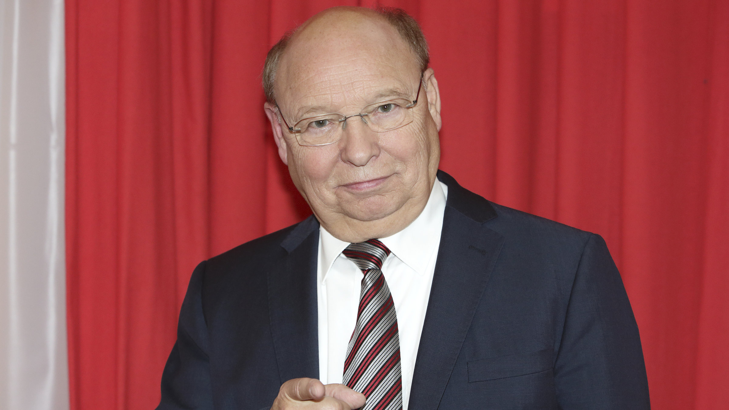 "Gernot Hassknecht: Jetzt wird's persönlich!": Gernot Hassknecht trägt einen dunkelblauen Anzug, ein weißesn Hemd und eine blau-rot-grau gestreifte Krawatte und schaut lächlend in die Kamera.