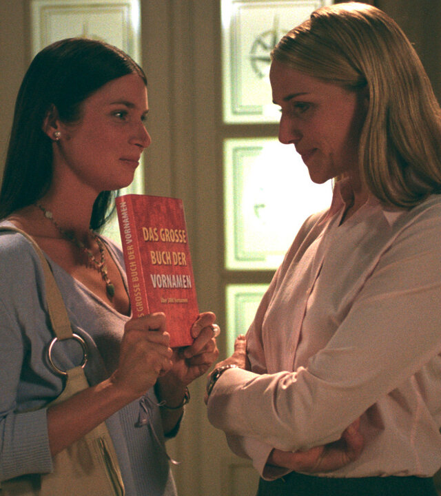 "Bianca - Wege zum Glück: Folge 30": Im Flur: Judith (Elisabeth Sutterlüty) zeigt Bianca (Tanja Wedhorn) ein Buch mit Vornamen für Babys. Bianca verschränkt ihre Arme und schaut auf das Buch.