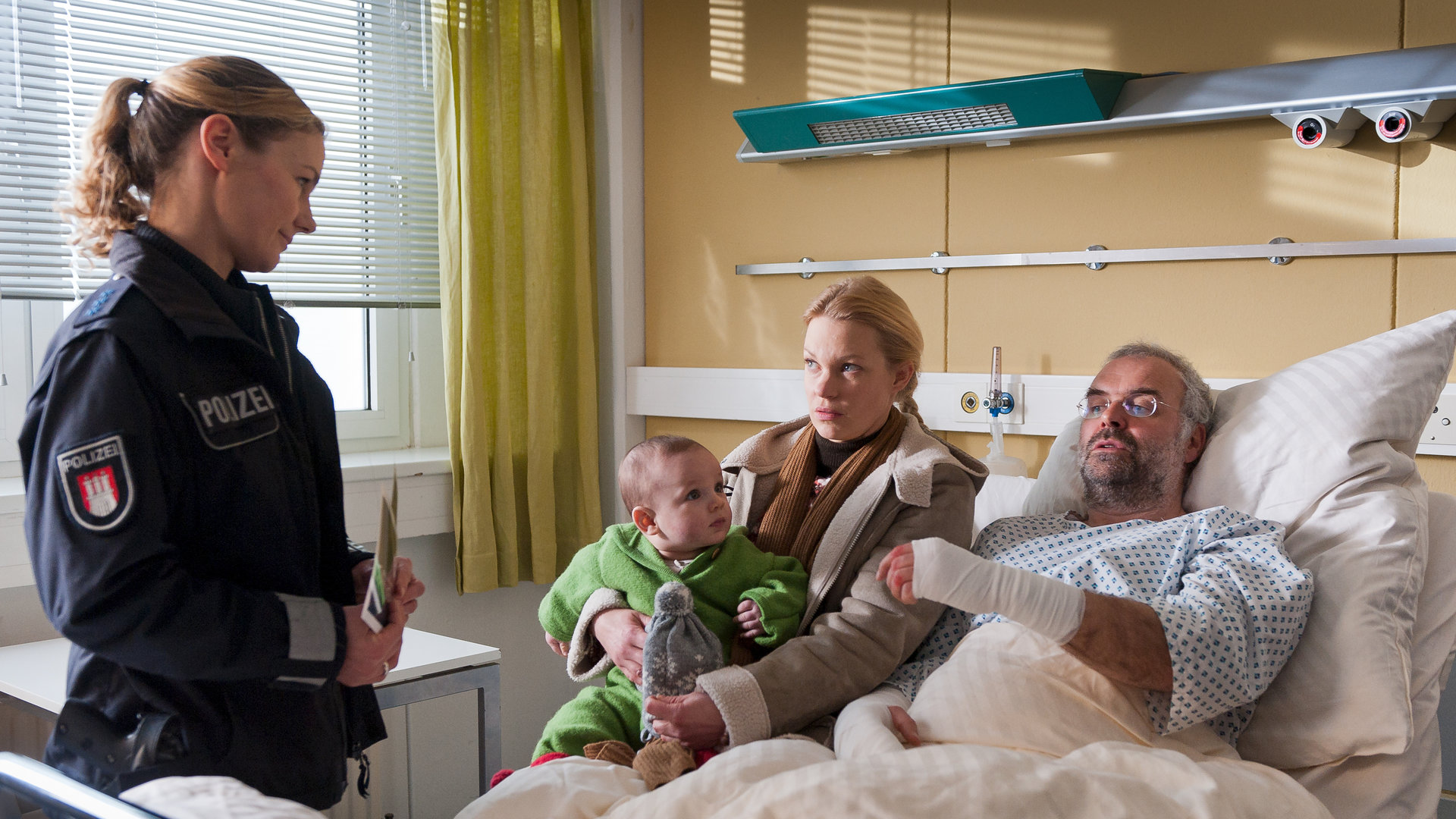 "Notruf Hafenkante - Leben daneben": Franzi (Rhea Harder) befragt Annik Karberg (Theresa Hübchen) und ihren Freund Enno Rüfken (Jophi Ries), der im Krankenbett liegt. Annik hat ein Kleinkind auf dem Schoß.