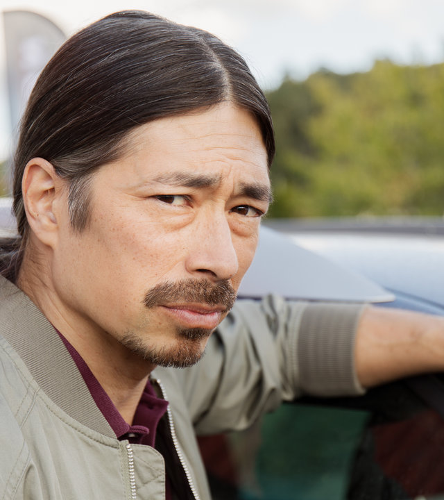 "Detective No. 24 - Suchen und Finden": Mats (Kristofer Kamiyasu) steht neben einem Auto. Sein linker Arm liegt auf dem Dach.