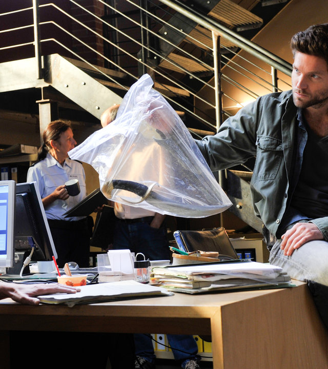 "SOKO Stuttgart - Major Mimis Ende": Selma Kirsch (Yve Burbach) sitzt an ihrem Schreibtisch und schaut angespannt zu Jo Stoll (Peter Ketnath), der auf der Tischkante sitzt und ihr einen Asservatenbeutel hinhält, in dem sich ein gebogener Dolch befindet. Im Hintergrund unterhalten sich eine Kollegin und ein Kollege.