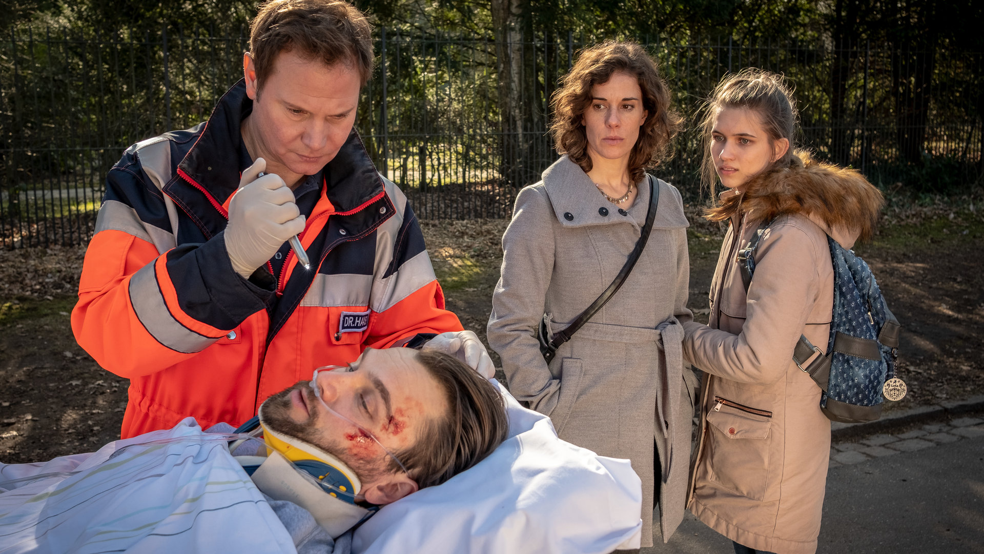 "Notruf Hafenkante - AB negativ": Notarzt Dr. Haase (Fabian Harloff) steht am Kopf einer Patiententrage, auf der Stefan Menzel (Matthias Kelle) liegt. Menzel hat Verletzungen im Gesicht. Dr. Haase leuchtet mit einer Pupillenleuchte auf Menzels rechtes Auge. Neben Dr. Haase stehen Annika (Anne Schäfer) und Tara Menzel (Anna-Lena Schwing), die besorgt zu Stefan Menzel sehen.
