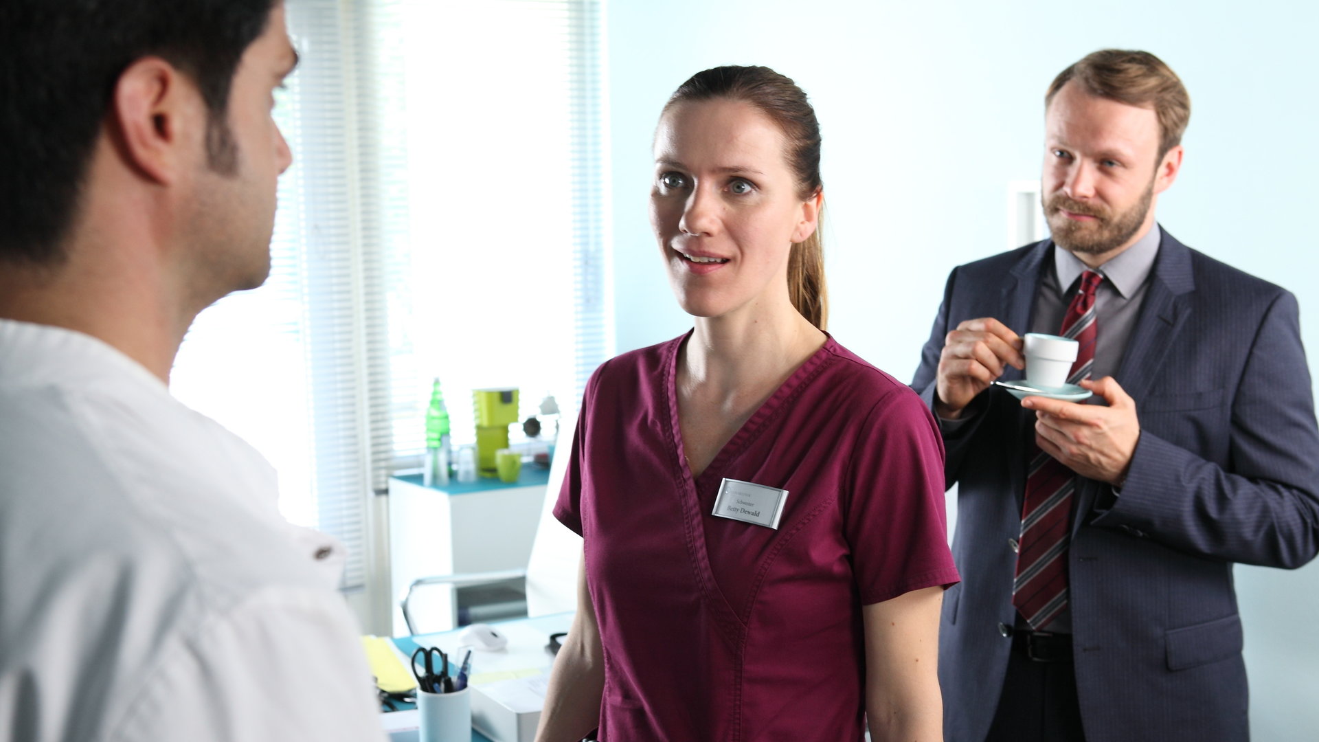 "Bettys Diagnose - Kein Tag wie jeder andere": Betty (Bettina Lamprecht) steht Dr. Behring (Maximilian Grill) in einem Zimmer gegenüber und schaut ihn ungläubig an. Hinter ihnen steht Jan Petersen (Matthias Ziesing), der gerade eine Tasse Kaffee trink und das Geschehen beobachtet.