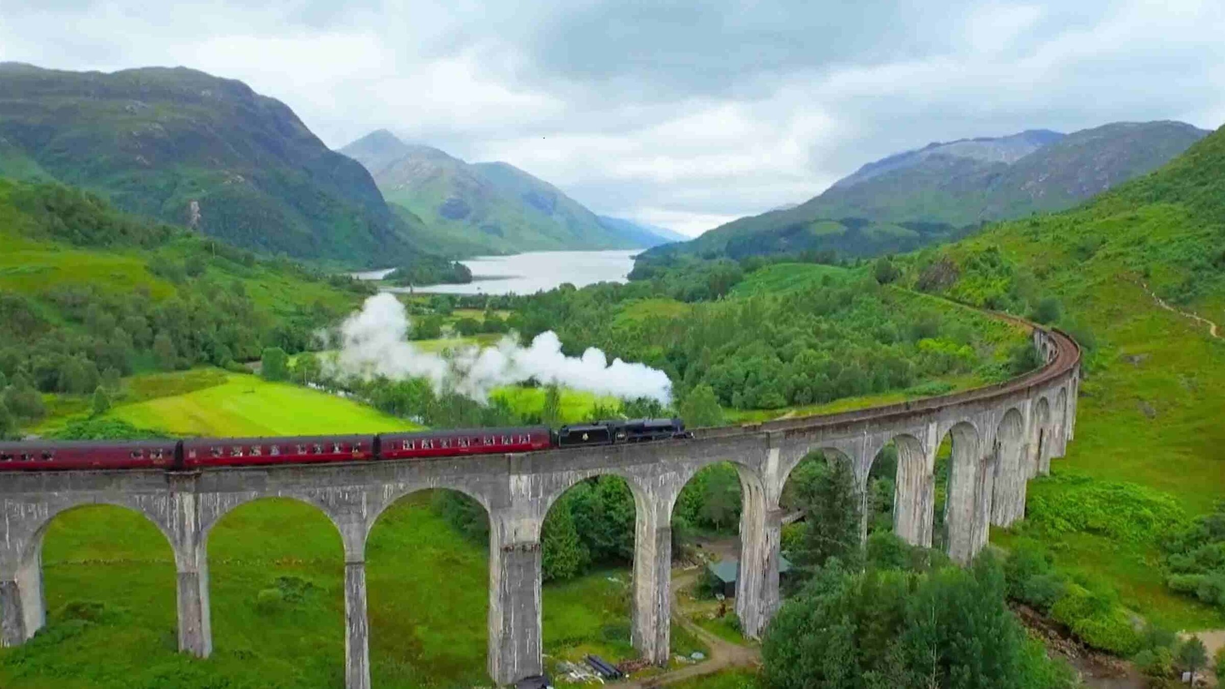 „Großbritanniens schönste Bahnstrecken - An Bord des Hogwarts Express": Eine rauchende Dampflok fährt auf einem bogenförmigen Eisenbahn-Viadukt über ein grünes Tal, im Hintergrund sieht man Berge und einen See